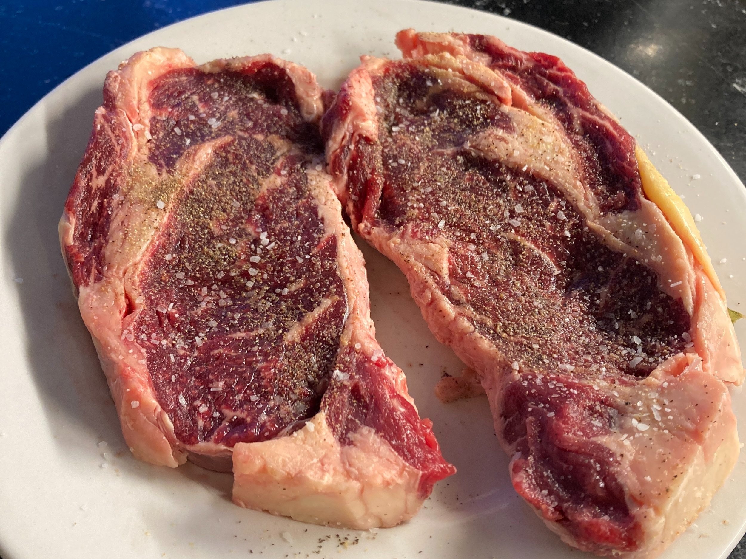 Two uncooked ribeye steaks seasoned with salt and pepper, placed on a white plate against a dark background, showcasing marbling and rich red color.